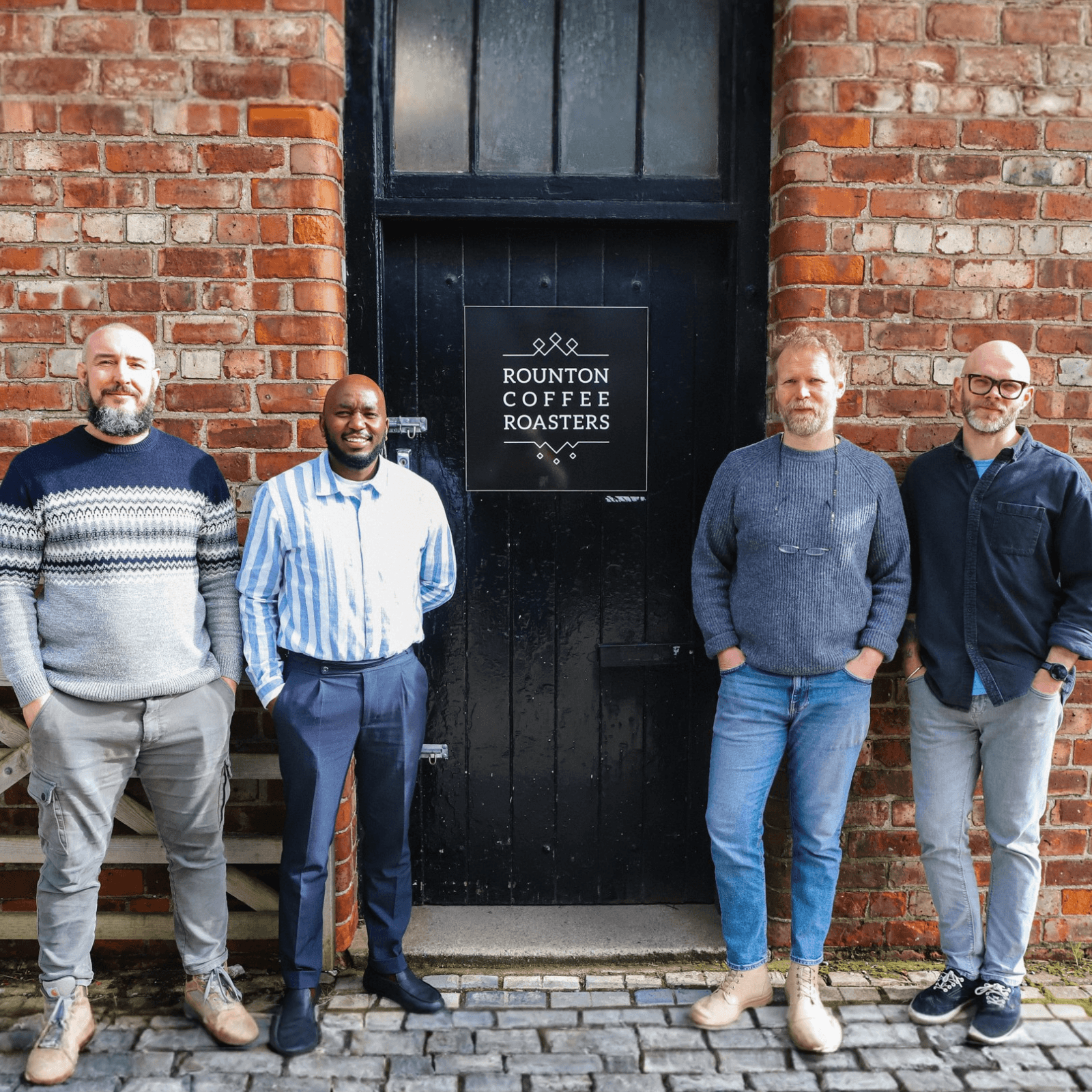 Four men standing in front of a brick building with 'Rounton Coffee Roasters' sign.