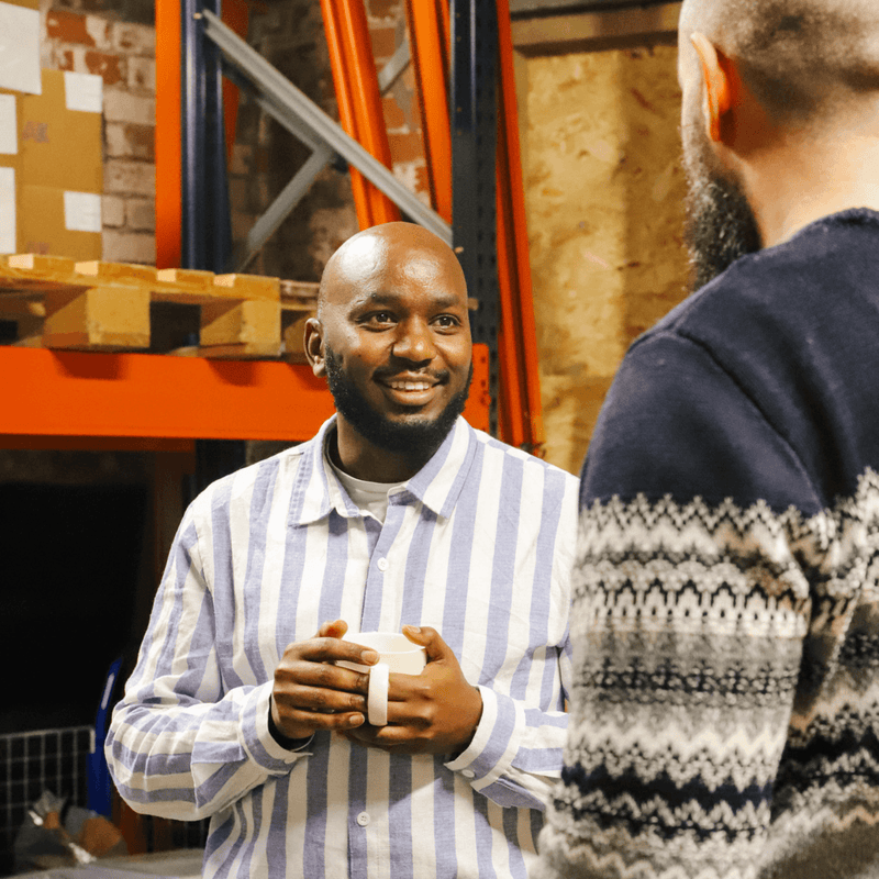 Two men talking in a warehouse with one holding a ceramic mug of coffee
